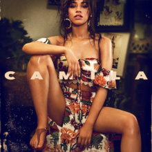 The cover image features Camila Cabello in foreground sitting on a small circular table wearing nature themed bra-like crop-top and long-skirt. In the background is a dim-lighted living-room. Above everything, in the middle, spread over whole width, in capitalised and bold format, is written the title 'CAMILA'.