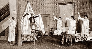 Stage scene in hotel bedroom with fully dressed man looking frantic, surrounded by young women in nightgowns