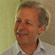 Alan, a White man with short, grey hair, is smiling away from the camera. He is wearing a blue & white, striped dress shirt and is in front of a concrete wall.