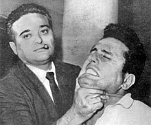 A black-and-white photograph showing a man wearing a jacket and tie with a short sigar in his mouth, on the left, holding up the chin of a man on the right, who appears to be in pain