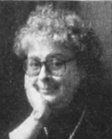 A white woman with curly grey hair, hand on cheek, wearing glasses