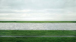 Uma fotografia do rio Reno inferior fluindo horizontalmente por campos verdes sob um céu nublado na Alemanha