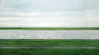 A photograph of the Lower Rhine river flowing horizontally through green fields under an overcast sky in Germany