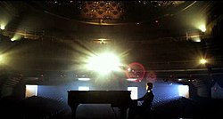 Bieber seen in an empty theater playing the piano while blue and yellow spot lighting is seen in the background.