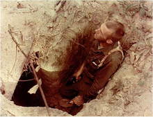 A soldier holding a pistol stands in a hole in the ground before the entrance to a tunnel