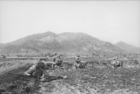 Soldados usando coletes à prova de balas e capacetes estão caídos em um campo aberto na base de uma grande colina com vegetação