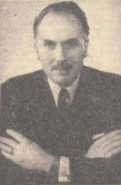 Foto de cabeça e ombros de um homem branco de meia-idade com cabelo escuro e bigode bem cuidado em uma suíte e gravata