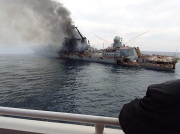A large warship lists heavily to her port side. Smoke billows from her bridge toward the photographer, who is at the rail of another vessel; the arm of someone else on that vessel is visible in the foreground.