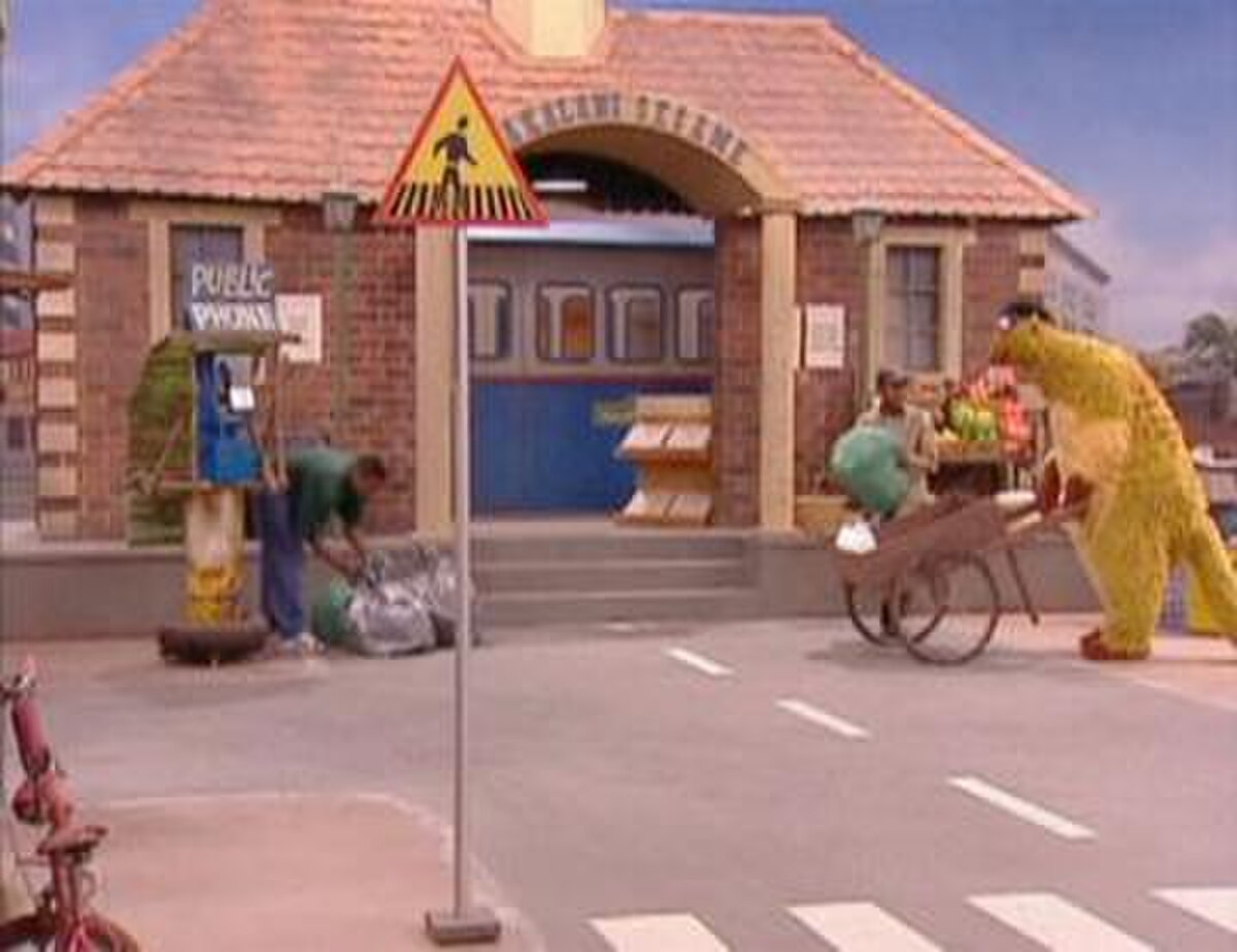 Television set， showing a large brick building on a city street; to the right， a large puppet is pushing a wheelbarrow and to the left， a man， next to a phone booth， is bending over several mail bags. Closer to the front of the image is a signpost， with a triangle that has a black figure and a yellow background， on top.