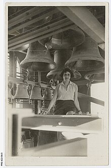 A woman with short, brown hair sitting on a wooden beam with large bells hanging in the background