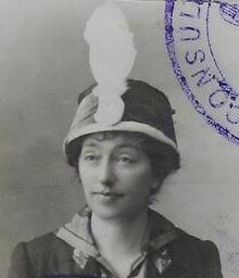 A black and white headshot photo of a woman who is looking forward to the left of the camera. She is wearing a hat with a large white feather attached to the front. The photo has part of a purple stamp in the top right corner with part of a word "CONSUL".