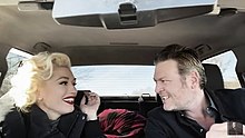 A color picture of singers Gwen Stefani and Blake Shelton, in the music video for their song "Nobody but You", sitting in a car in front of a green screen; Shelton is driving.