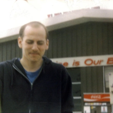 Photo of Adam Demirjian infront of a black gas station with a white roof. He is wearing a black hoodie which is slightly open, revealing a blue t-shift. He appears to be walking away from the gas station. His eyes are closed.