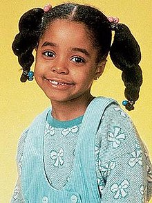 A young African American girl approximately aged five or six smiling at the camera. Dressed in a blue jumper and overalls, her long black hair is worn in a pair of pig tails that protrude from opposite sides of her head.