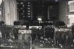 Police at the Broadwater Farm riot, with long shields, NATO helmets, and fire-resistant overalls, but no body armour Broadwater Farm riot, 1985.jpg