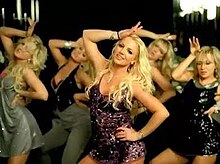 Image of five blond women dancing in a big bathroom. The woman in the front is smiling and wearing a purple satin dress.