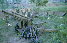 Im Vordergrund steht ein beschädigtes Artilleriegeschütz, darauf ruhen mehrere Gewehre. Im Hintergrund stehen im Hintergrund mehrere kaukasische Soldaten neben einem Hootchie.
