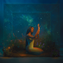 A woman holds both hands up towards a light shining on her; she sits in an underwater cube that reflects the cosmos.