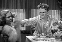 Cagney, in striped pajamas, looks angry as he reaches across a breakfast table with the grapefruit in his hand.