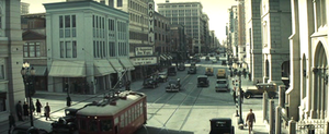 Elevated daytime view of a busy city crossroads, looking down one street along which a series of tall buildings recede into the distance. Pedestrians line the sidewalks, cars litter the streets; a bright red electric streetcar features in the foreground