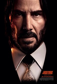 A closeup of John Wick, wearing a hourglass as a "tie", with bullets flowing into the lower bulb inside the hourglass.