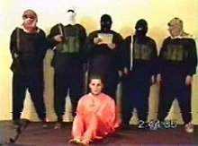 A photo of a young man with a beard and overgrown hair, dressed in orange, seated on the floor as five men stand over him. The men wear black, face coverings, face masks and military vests. The man in the center holds a plaque with unreadable text. The photo is very blurry.