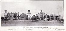 Une photographie en noir et blanc montrant les bâtiments construits/conçus par Stanley.  De gauche à droite, ce sont l'école technique, la petite salle, la tour de l'horloge, la grande salle et la maison du secrétaire.  Ils sont vus de l'autre côté de la route, avec des clôtures devant les bâtiments.