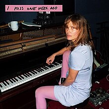 A colored photograph of Oh Land sitting at a piano in a dark studio room.