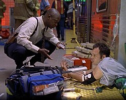 A man wearing a white shirt and blank pants kneels and speaks to a man in a blue shirt who is pinned between a subway train car and a platform. Emergency equipment lies in front of them, while the obscured figures of firefighters and emergency personnel stand behind them.