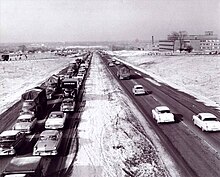 Une photo en noir et blanc montre une autoroute à quatre voies divisée par un terre-plein en herbe.  Dans les voies venant en sens inverse, le trafic est congestionné au loin.  À quelques exceptions près, la 401 est entourée de terres agricoles.