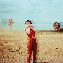 A woman stands in the middle of a field, covering her face with her hands