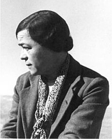 Photograph of a woman in a formal jacket and tied-up hair, facing slightly away from the camera