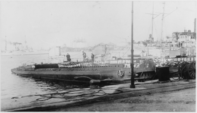 Submarines are docked in a port side-by-side. A battleship can be seen in the background.