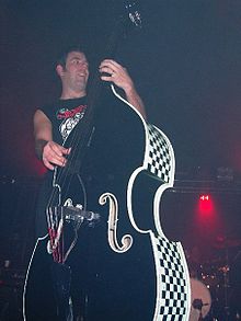 Owen is playing his double bass guitar. He is behind and obscured by it. His left hand is high on the neck and he wears a gold ring on his third finger. His right hand is plucking the strings while he is looking to his left. A red stage light is visible behind him. He partly obscures a drum kit. The side of his double bass has a black and white chequered design.