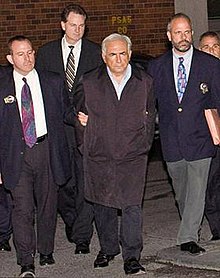 A white-haired man in a black overcoat and dress shirt with his hands behind his back at the center of a small group of men walking toward the camera. The two men on either side are wearing jackets with gold badges clipped to the lapels and ties. They are holding the arms of the man in the center. A fourth man, also in a jacket and tie, is visible in the rear.