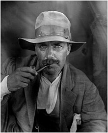 Calder as a young man, in an outdoor hat and dirty clothing, smoking a pipe.