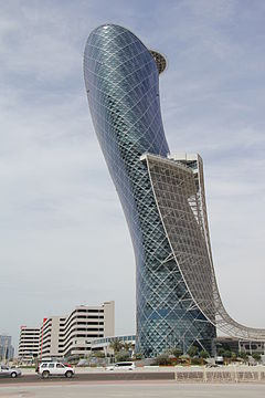 фотография высокого, стеклянного, несколько цилиндрического здания, которое начинается прямо и наклоняется влево