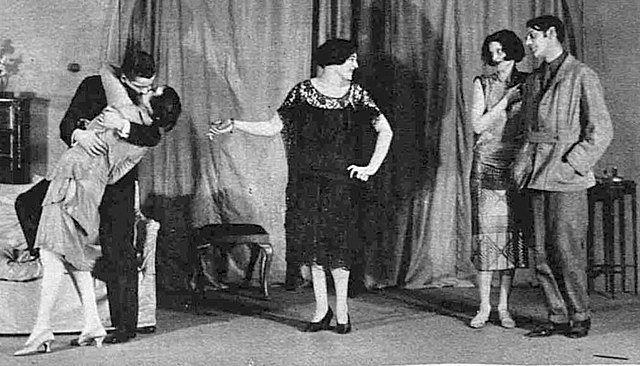 stage scene, indoor setting, a woman standing centre, with to her right an embracing couple and to her left another couple, looking on benevolently