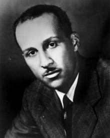 Studio portrait photograph of a young black man, in a formal jacket and tie, with short hair and a short, neat moustache.