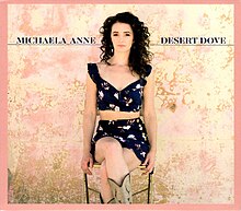 The cover features a woman sitting on a chair, wearing a navy blue top and skirt with flower patterns and beige cowboy boots, with a stained peach wall behind her. The artist's name and album title appear on the left and right of the artist, colored in navy blue.
