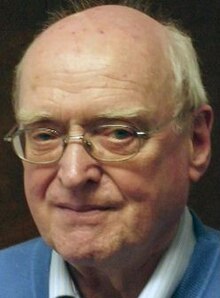 Colour photo of elderly, half-smiling, bald and clean-shaven man with wire-framed spectacles