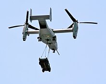 A MV-22 with its rotors up to vertical with a HMMWV vehicle hanging by two sling wires.