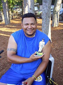 Mies, jolla on siniset hihattomat t-paidat ja siniset shortsit, hymyilee pitäessään itsensä valkoisessa baseball-uniformissa kuvaavaa bobbleheadia.