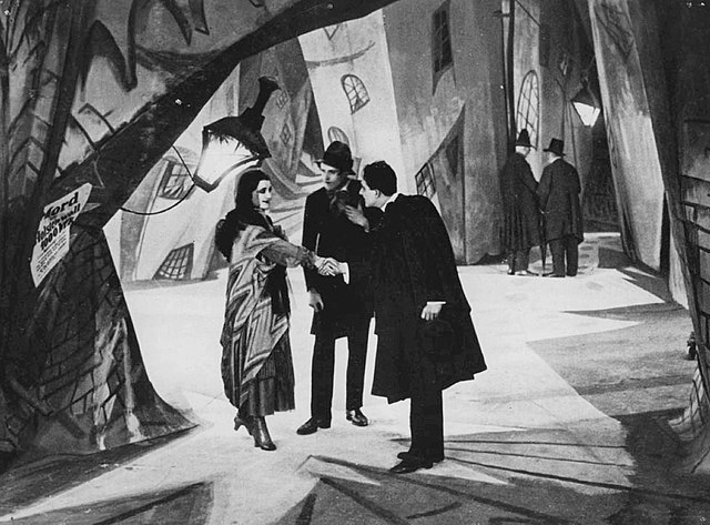 A man shakes hands with a woman, while another man looks on between them. The three figures stand in the iddle of a city street, with brick walls in twisted and distorted shapes, and shadows and streaks of light painted onto the walls and ground.