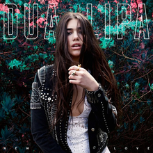Dua Lipa in a black jacket posing in front of a floral background.