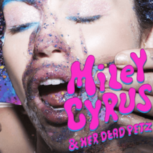 Cyrus emerging from a pool of glitter, smearing glitter with the same colors on her cheek.