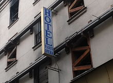 Enseigne verticale en lettres majuscules bleues sur fond blanc, le mot HOTEL écrit verticalement et en-dessous, les mots PARIS et OPERA sur deux lignes horizontales, devant des fenêtres soutenues par des tasseaux.