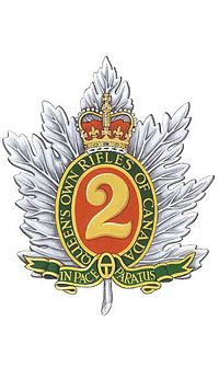 Kolorowy obraz odznaki wojskowej składającej się ze srebrnego liścia klonu ze złotą cyfrą „2” pośrodku na czerwonym tle, otoczonym zielonym, owalnym listello ze złotymi krawędziami, na którym widnieje „Queen's Own Rifles of Canada” napis wykonany dużymi złotymi literami zwieńczony naturalną koroną królewską oraz, na łodydze liścia klonu, zielony pasek obszyty złotem z napisem `` In Pace Paratus '' wielkimi literami w kolorze złotym