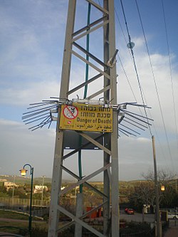 אזהרה תלת-לשונית מפני התחשמלות, יחד עם גדר שמונעת טיפוס, על עמוד חשמל של חברת החשמל לישראל.