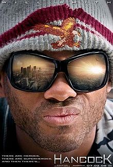 Close portrait of a man with stubble on his face and large sunglasses in which the reflection of a city's landscape, awash in sunlight, can be seen. The man is wearing a gray knitted cap with a dark red rim; stitched in the front of the cap is a orange-tinted bald eagle with its wings spread. In the lower right corner of the poster is the name "HANCOCK".